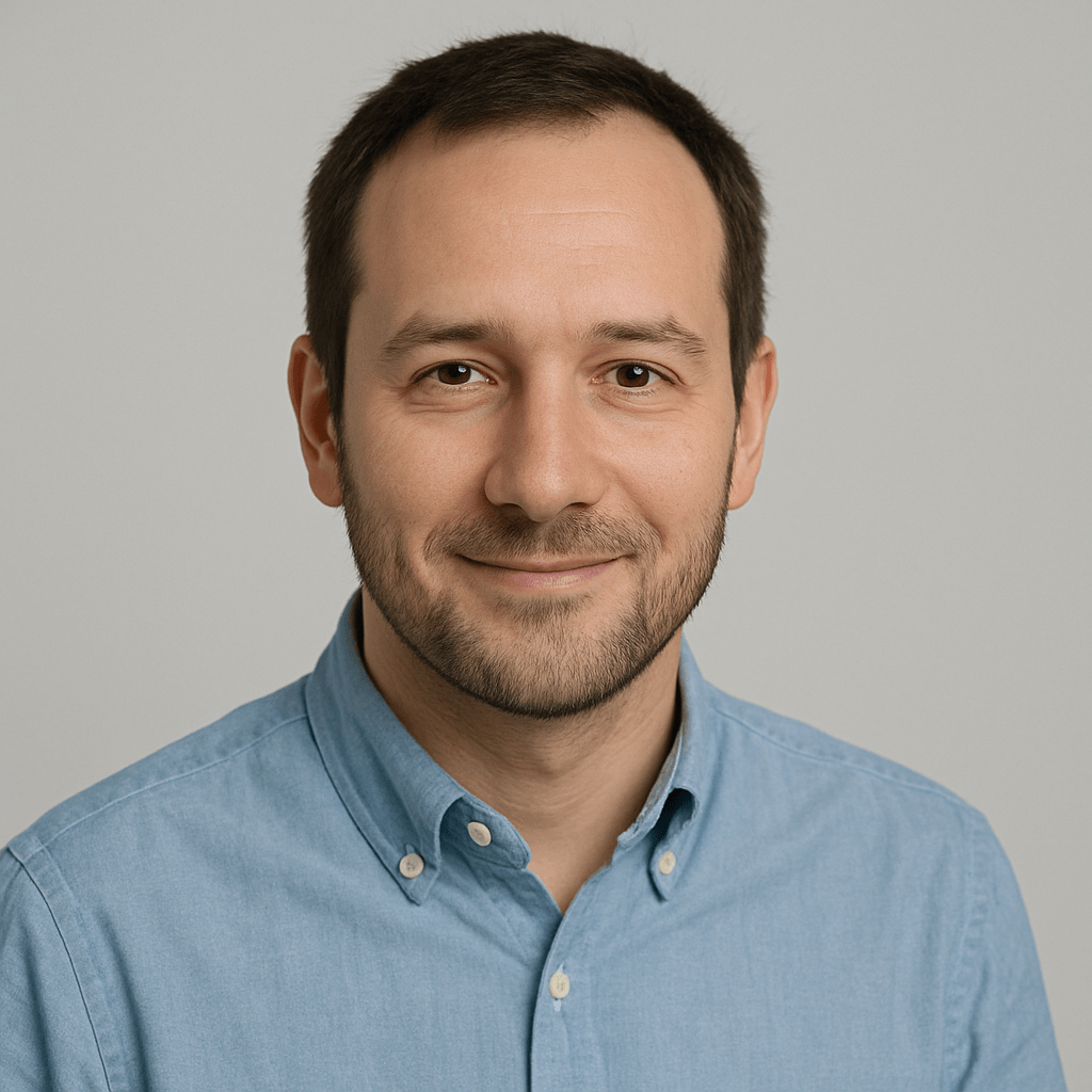 Porträt eines lächelnden Mannes mit kurzem Bart und blauem Hemd vor blau-grauem Hintergrund.