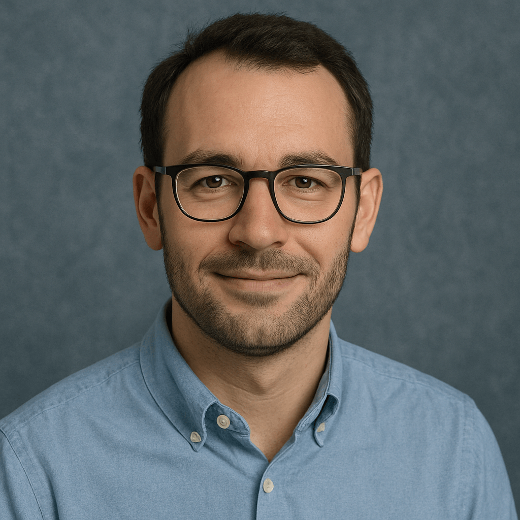 Porträt eines Mannes mit dunkler Brille, kurzem Bart und hellblauem Hemd vor blau-grauem Hintergrund.